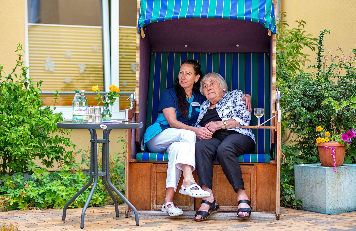 Pflegekraft umarmt betreute Person im Strandkorb